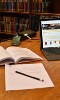 library books in background with a laptop, paper, pencil and open book on a table in the foreground